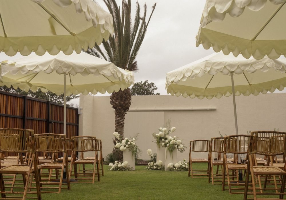 The ceremony set on the lawn: bamboo chairs, white florals, a palm and scalloped umbrellas