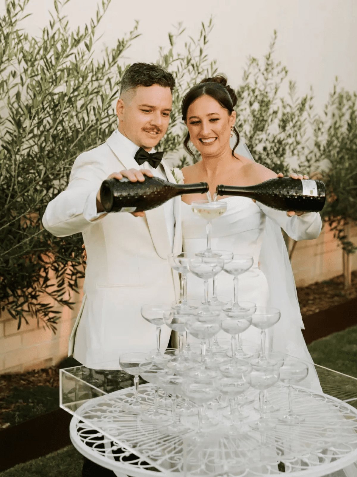 A couple in white pouring champagne into a coupe tower among olive trees