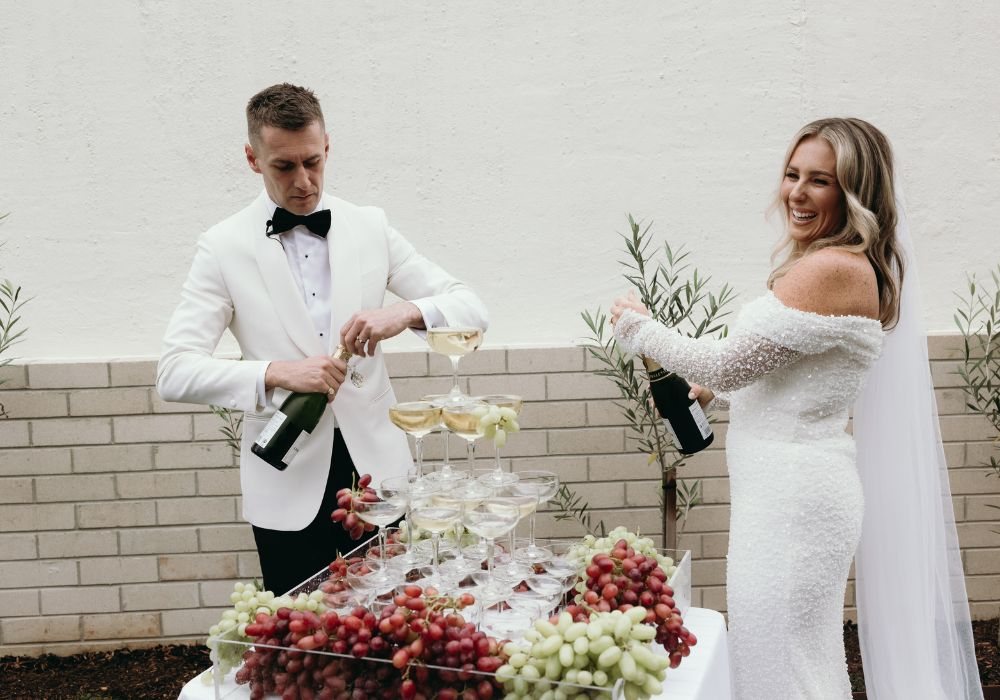 Bride and groom opening champagne beside a coupe tower against the white wall