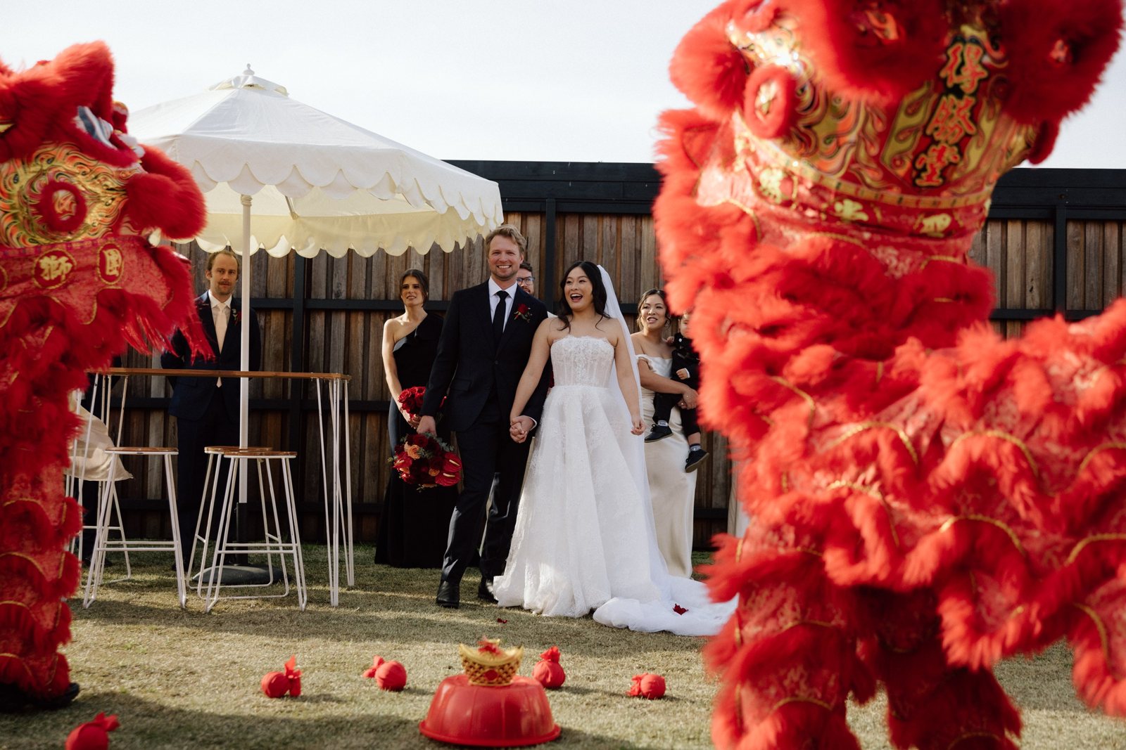 A couple walking onto the lawn between two red lion dancers, umbrellas behind