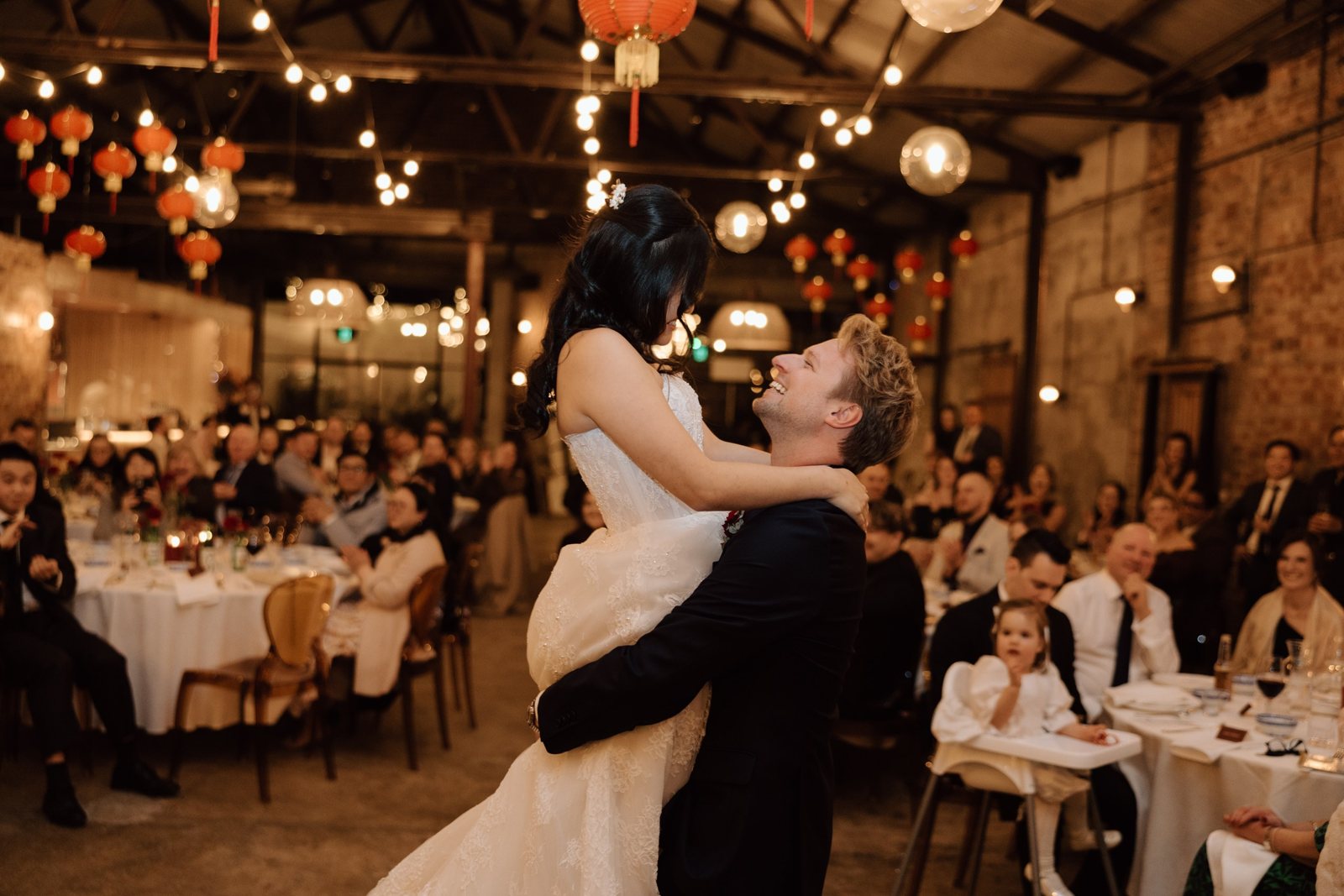 The groom lifting the bride in the Truck Room, guests at round tables around them