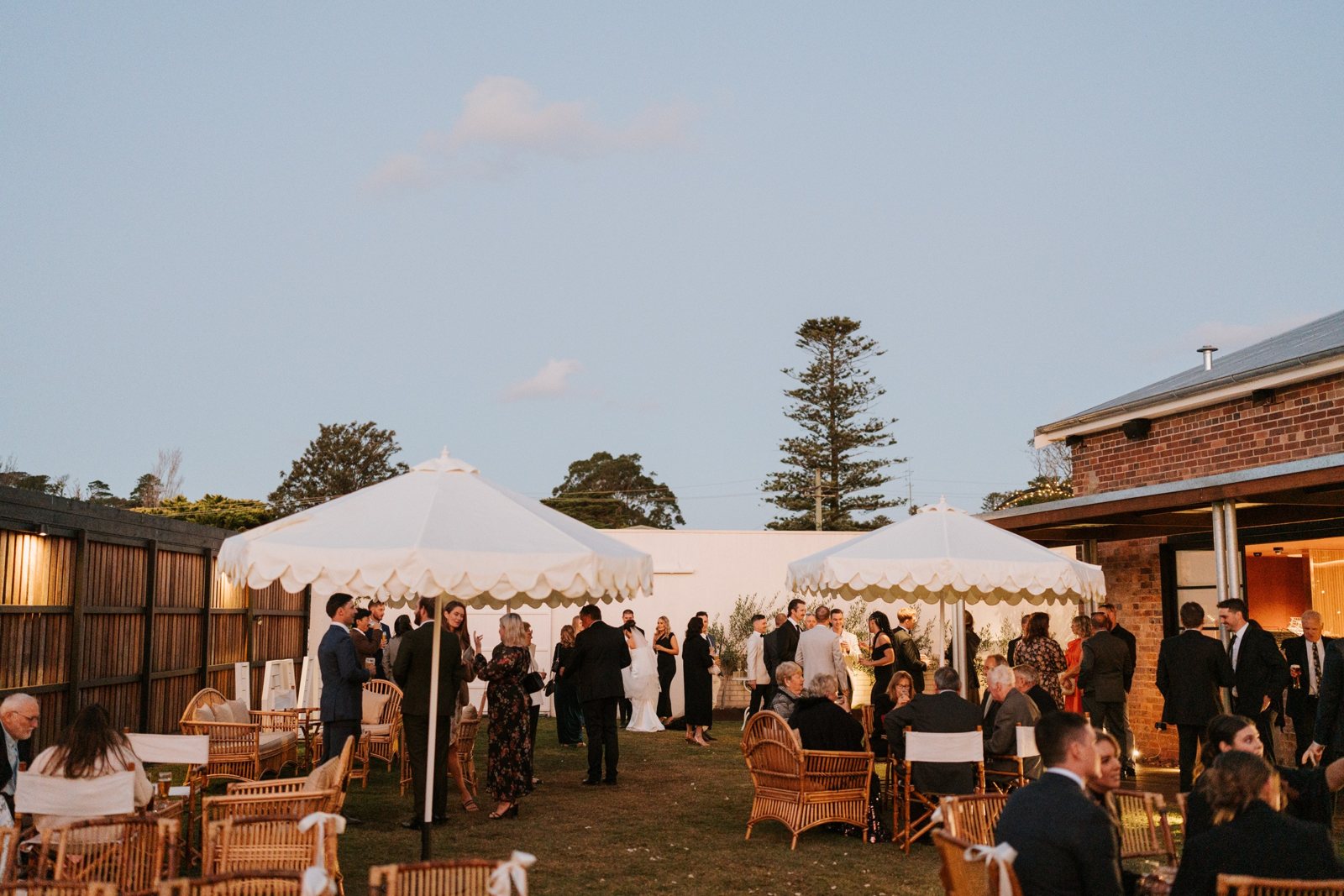 Guests at dusk under scalloped umbrellas on the lawn beside the brick building